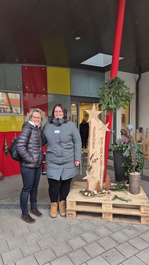 BLP-Vorstandsmitglied Gabriela Hirschhofer sowie Jasmin Tinhof, Standortleiterin von pro mente in Mattersburg, stehen lächelnd vor einem Eingang; daneben ein Weihnachtsdekor auf Paletten mit Holzstele "Schöne Weihnachtszeit", Sternen und Tannengrün.