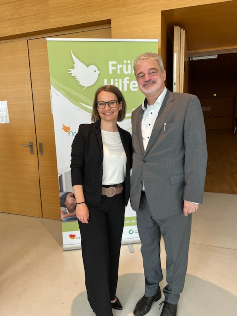 Zwei Personen stehen lächelnd bei einer Veranstaltung vor einem Roll-up-Banner von „Frühe Hilfen“. Die Frau trägt ein schwarzes Business-Outfit, der Mann einen grauen Anzug. Beide posieren in einem modernen Innenraum nebeneinander.