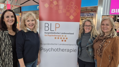 Vier Frauen stehen nebeneinander und lächeln in die Kamera. Hinter ihnen steht ein Roll-Up mit der Aufschrift „Burgenländischer Landesverband für Psychotherapie“. Die Frauen tragen Freizeitkleidung und stehen in einem Einkaufszentrum.
