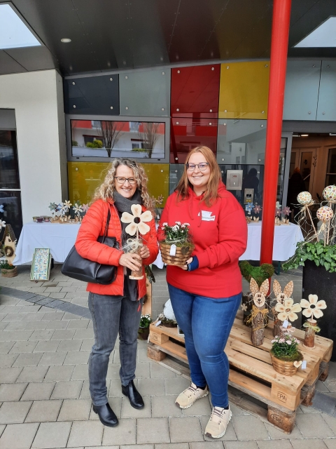 Zwei lächelnde Frauen auf einem Frühlingsmarkt vor einem Gebäude mit bunten Glasplatten. Beide in roten Jacken, mit Blumendeko in den Händen.