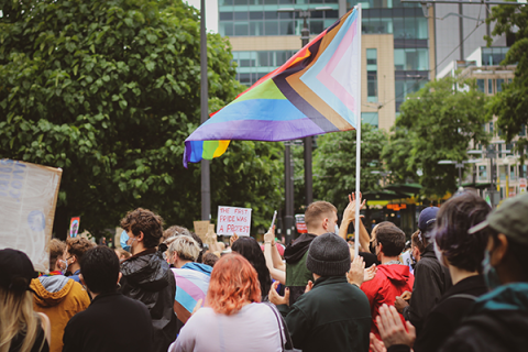 Bild einer Pride-Kundgebung. Zu sehen ist eine größere Menschenansammlung, es werden Schilder und eine Progress-Pride-Flagge in die Luft gehalten. 
