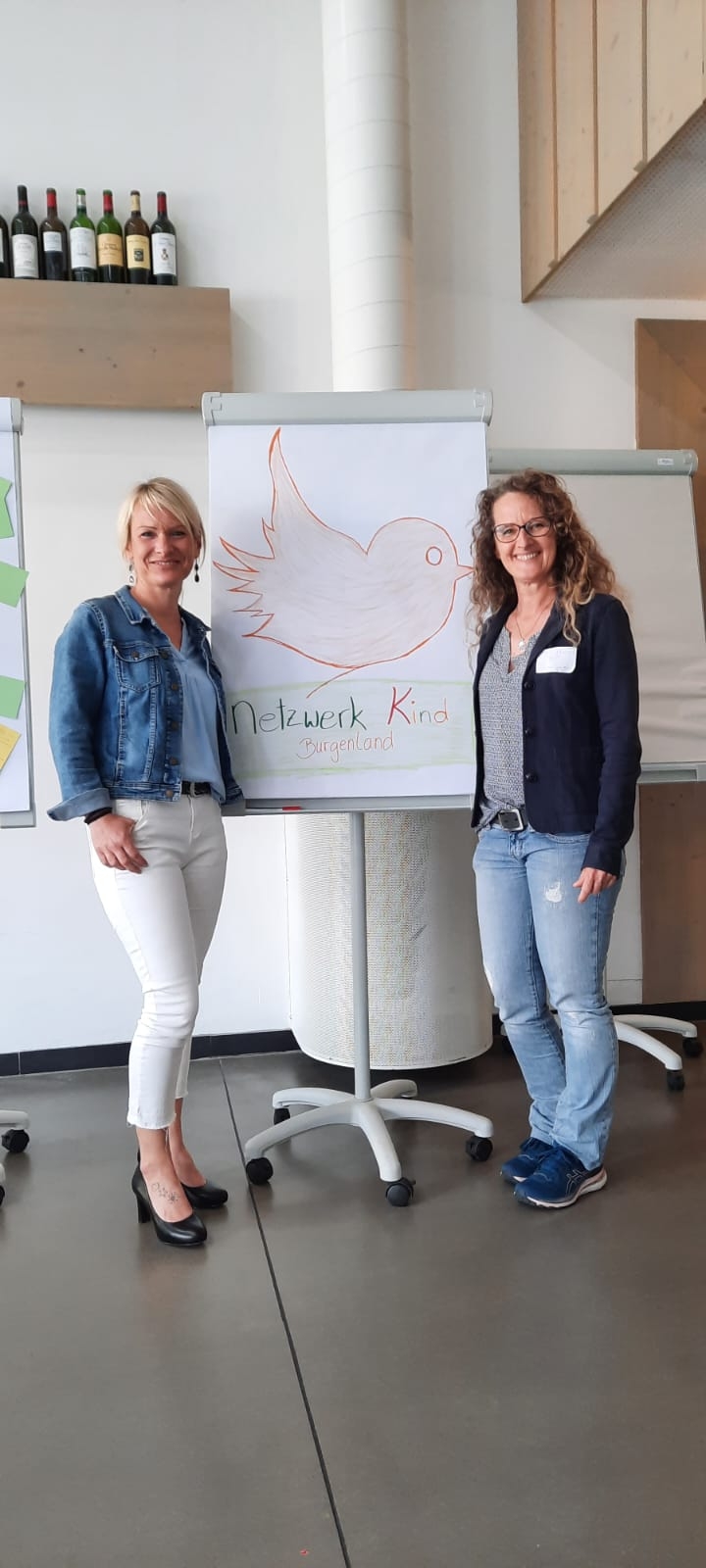 Organisator:innen des runden Tisches von "Netzwerk Kind Burgenland" stehen lächelnd links und rechts neben einem Flipchart. Au dem Flipchart ist ein roter Vogel zu sehen mit dem Text "Netzwerk Kind Burgenland".