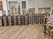 Ausstellung handgefertigter weihnachtlicher Holzdekorationen in einem Innenraum, mit beleuchteten Holzrahmen, Weihnachtskugeln, kleinen Figuren, Sternen, Miniaturhäusern und weiteren dekorativen Elementen, die auf Tischen und am Boden präsentiert werden.