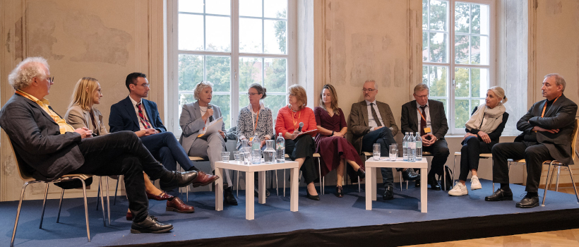 Podiumsdiskussion mit mehreren Teilnehmer:innen in einem hellen Raum, sitzend im Halbkreis vor großen Fenstern, Gesprächssituation mit Mikrofonen und Tischen mit Wasserflaschen.